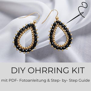 Puede incluir: Un par de pendientes en forma de lágrima con cuentas negras facetadas y detalles en tono dorado con ganchos dorados. El texto DIY OHRRING KIT mit PDF- Fotoanleitung & Step- by- Step Guide aparece impreso bajo los pendientes.