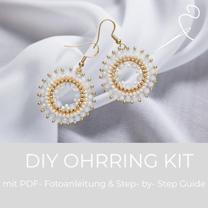 May include: A pair of handmade earrings with a circular design. The earrings feature a gold-colored metal hook and are adorned with white and gold beads. The text "DIY OHRRING KIT" is displayed at the bottom of the image.