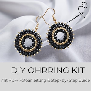 Puede incluir: Un par de pendientes circulares con cuentas negras facetadas y detalles en tono dorado sobre ganchos de color oro. El texto DIY OHRRING KIT mit PDF- Fotoanleitung & Step- by- Step Guide está impreso debajo de los pendientes.