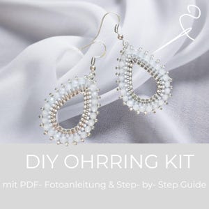 Pode incluir: Um par de brincos em forma de gota com contas brancas e prateadas metálicas dispostas num padrão circular. Os brincos estão suspensos em ganchos de metal prateado. O texto DIY OHRRING KIT mit PDF- Fotoanleitung & Step- by- Step Guide é visível.