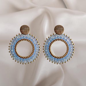 Puede incluir: Un par de pendientes de aro con un diseño de cuentas azul claro. Los pendientes tienen un marco de metal dorado y una pequeña parte superior redonda y texturizada de color dorado. Se muestran sobre un fondo blanco y sedoso.