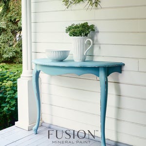 Puede incluir: Una mesa consola de media luna de color azul claro con pintura blanca desgastada en las patas. La mesa est&aacute; situada en un porche de madera con una pared blanca en el fondo. El texto "FUSION MINERAL PAINT paint it beautiful." es visible en la esquina inferior derecha de la imagen.