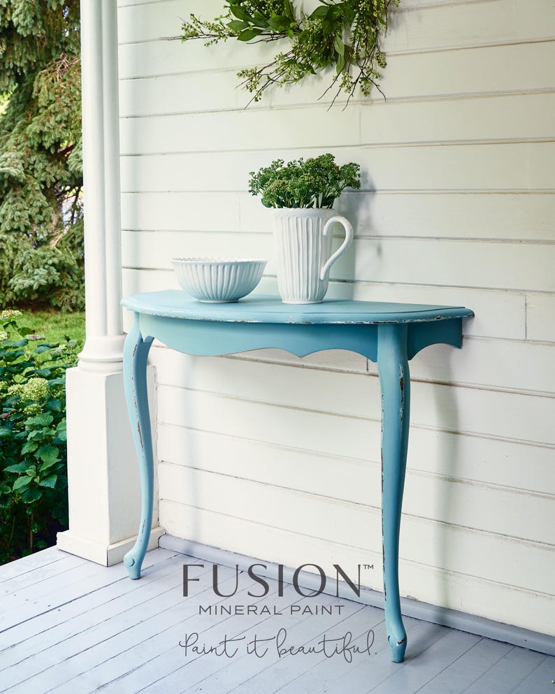 Puede incluir: Una mesa consola de media luna de color azul claro con pintura blanca desgastada en las patas. La mesa est&aacute; situada en un porche de madera con una pared blanca en el fondo. El texto "FUSION MINERAL PAINT paint it beautiful." es visible en la esquina inferior derecha de la imagen.