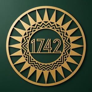 Può includere: Una placca circolare color oro con il numero 1742 al centro. Il design presenta un motivo a raggiera con punte triangolari che si irradiano verso l'esterno e un bordo decorativo a zigzag che circonda la visualizzazione numerica centrale.