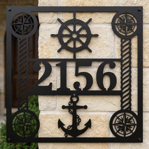 May include: A square black metal house number sign featuring the digits 2156 in the center. The design includes a ship's wheel at the top, an anchor at the bottom, four compass roses in the corners, and two vertical rope-patterned side pillars.