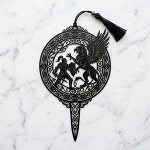 May include: A black laser-cut cake topper featuring a circular frame with intricate filigree patterns. Inside the circle, a knight in armour stands with a sword and shield facing a griffin with spread wings. A black tassel is attached to the top.