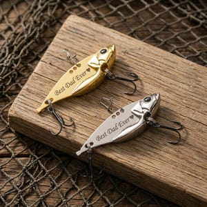 Puede incluir: Dos señuelos de pesca, uno dorado y otro plateado, descansan sobre una superficie de madera. Cada señuelo tiene forma de pez con anzuelos y el texto grabado "Best Dad Ever" con un corazón. Un fondo de red de pesca.