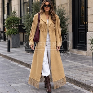 May include: A long tan coat featuring fringe detailing along the sleeves and hemline, worn over white trousers. The coat has a notched collar and a button-front closure. A dark burgundy shoulder bag is carried on one side of the garment.