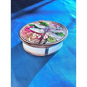 May include: An oval-shaped trinket box with a white base and a gold-colored rim. The lid features a stained-glass design of a dragonfly and pink and purple flowers. The box is set against a blue background.
