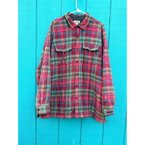May include: A long-sleeved, button-down flannel shirt in a classic plaid pattern. The shirt features a mix of red, green, and blue hues, with two chest pockets and a collared neckline. The shirt is hanging on a white hanger against a turquoise background.