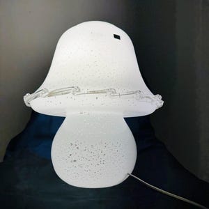 Può includere: Una lampada bianca a forma di fungo con un design maculato. La lampada ha una base arrotondata e una parte superiore svasata, con una catena decorativa di vetro sul bordo. La lampada è illuminata e un cavo di alimentazione si estende dalla base.