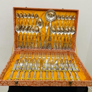 Puede incluir: Un juego de cubiertos antiguos en un estuche decorativo. El juego incluye cucharas, tenedores y cuchillos, todos con mangos ornamentados. El estuche está forrado con tela amarilla y tiene un estampado floral en el exterior.