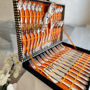 Puede incluir: Un juego de cubiertos de plata exhibido en un estuche abierto a cuadros blancos y negros con interior naranja. El juego incluye cucharas, tenedores y cuchillos, asegurados con cintas naranjas. El estuche está abierto, revelando la cubertería cuidadosamente dispuesta.