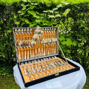 Könnte beinhalten: Ein silberfarbenes Besteckset in einem schwarzen Koffer mit Schachbrettmuster. Das Set enthält Löffel, Gabeln und Messer, die auf einem orangefarbenen Hintergrund angeordnet sind. Der Koffer ist geöffnet und zeigt das komplette Set auf einer weißen Tischdecke.