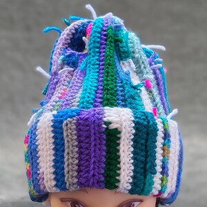 May include: A colorful, handmade crochet hat with a striped pattern in shades of blue, purple, white, and green. The hat has a textured appearance with small, protruding yarn ends and pink accents. The hat is displayed on a mannequin head.