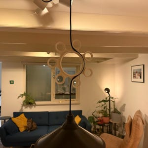 Könnte beinhalten: Eine moderne, weiße, geometrische Pendelleuchte mit einem schwarzen Kabel. Die Leuchte hat ein kreisförmiges Design mit mehreren kleineren kreisförmigen Aussparungen. Die Leuchte hängt in einem Wohnzimmer mit einem blauen Sofa und gelben Kissen.