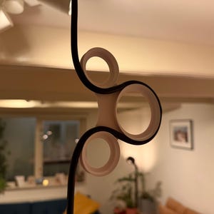 Può includere: Un moderno lampadario astratto con un cavo nero ed elementi circolari bianchi. Il lampadario ha un design scultoreo unico con più anelli interconnessi. Lo sfondo è un interno dai toni caldi.