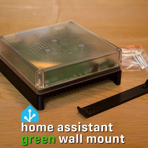 May include: A Home Assistant Green device featuring a clear, semi-transparent top cover and a black base, accompanied by a separate black mounting bracket and a small plastic bag containing four silver metal screws for installation purposes.