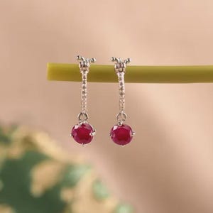 Pode incluir: Um par de brincos de argola em prata com uma banda fina com pequenas pedras incrustadas e uma única pedra preciosa redonda de cor vermelho-profundo pendurada na base de cada argola. Os brincos estão suspensos num caule verde.