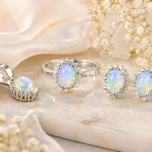 May include: A jewelry set consisting of a silver-toned ring, a pair of stud earrings, and a pendant. Each piece features a central oval-shaped iridescent opal stone surrounded by a halo of small, clear, round-cut stones set in silver metal.