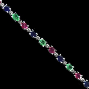 Puede incluir: Una pulsera de tenis con piedras preciosas multicolores que presenta un patrón repetitivo de zafiros azules, esmeraldas verdes y rubíes rojos de corte ovalado. Cada gema está separada por pequeños diamantes blancos redondos en una banda de metal plateado.
