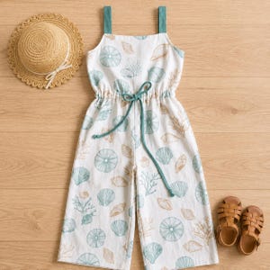 Op de afbeelding: Een witte mouwloze jumpsuit met een print van blauwgroene en beige schelpen en koraal, met blauwgroene schouderbandjes en een trekkoord in de taille, vergezeld door een beige strooien hoed met wit lint en een paar bruine leren sandalen.