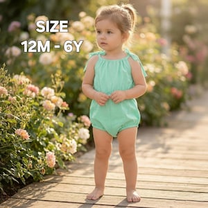 May include: A young child wearing a mint green romper with tie straps. The romper is a one-piece garment with a gathered neckline and a bubble-style bottom. The text "SIZE 12M - 6Y" is in the upper left corner. The child is standing on a wooden surface.
