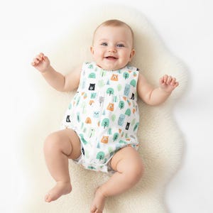 Pode incluir: Um bebé a usar um macacão branco sem mangas com um padrão colorido de ursos e árvores. O macacão apresenta ursos e árvores pretos, laranja e verde-azulado. O bebé está deitado numa superfície branca e fofa e está a sorrir.