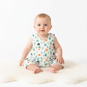 May include: A sleeveless baby romper featuring a white base with a repeating pattern of small brown bears, black bears, and stylised green and blue trees. The garment has a V-neckline and a loose, comfortable fit for an infant to wear.