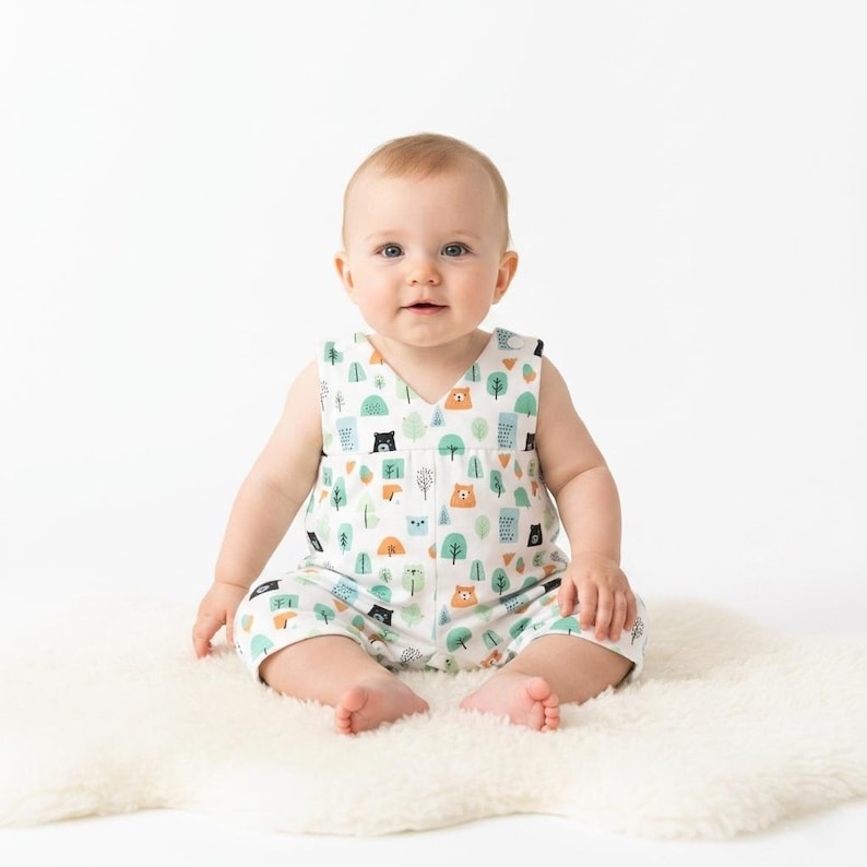 May include: A sleeveless baby romper featuring a white base with a repeating pattern of small brown bears, black bears, and stylised green and blue trees. The garment has a V-neckline and a loose, comfortable fit for an infant to wear.