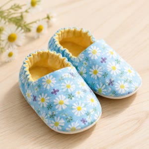 May include: A pair of baby shoes featuring a light blue fabric exterior with a repeating pattern of white daisies, small green leaves, and tiny purple flowers. The shoes have a soft, yellow interior lining and an elasticized opening for a snug fit.