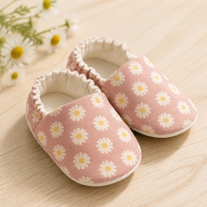 May include: A pair of soft baby shoes featuring a dusty pink fabric base with a repeating white and yellow daisy flower pattern. The shoes have an elasticized gathered opening and a simple slip-on design with a rounded toe and white interior lining.