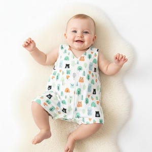 May include: A sleeveless baby romper featuring a white base with a repeating pattern of stylised brown bears, black bears, and various green and blue trees. The garment has a V-neckline and a relaxed fit, designed for comfort and ease of movement.