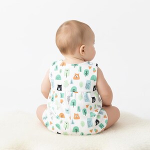 May include: A sleeveless white romper featuring a repeating pattern of stylised brown bears, black bears, and various green and orange trees. The garment has a gathered waistline and a simple round neckline, showcasing a playful forest-themed print.