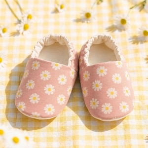 May include: A pair of soft baby slippers in a dusty pink color featuring a repeating white daisy flower pattern with yellow centers. The slippers have a rounded toe design, a gathered elasticized opening, and a light-colored interior lining.