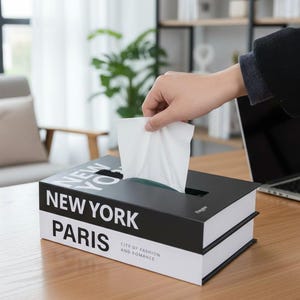 Op de afbeelding: Een zwart-witte tissuebox ontworpen als een boek, met de woorden "NEW YORK" en "PARIS" op de zijkant gedrukt. De doos staat op een houten tafel en een hand reikt naar binnen om een tissue te pakken. De tekst "CITY OF FASHION AND ROMANCE" staat ook op de doos.