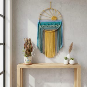 May include: A handcrafted wall hanging with a sun and ocean design. The circular frame features a yellow sun, blue and turquoise waves, and vertical strands in yellow, blue, and gray. It is displayed on a light wooden table.