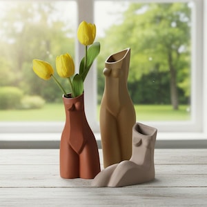 Pode incluir: Três vasos de cerâmica em forma de corpo feminino. Um é terracota, segurando tulipas amarelas. Os outros dois vasos são bege e taupe. Os vasos são exibidos em uma superfície de madeira branca em frente a uma janela.