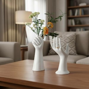 Puede incluir: Dos jarrones de cerámica blanca con forma de manos, uno con el puño cerrado sosteniendo flores, el otro con la mano abierta con anillos. Las flores son verdes y naranjas. Los jarrones están sobre una mesa de madera.