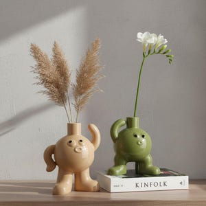 May include: Two whimsical ceramic vases, one beige and one green, each with a unique anthropomorphic design. The beige vase holds dried pampas grass, while the green vase displays a white flower. The vases are on a wooden surface.