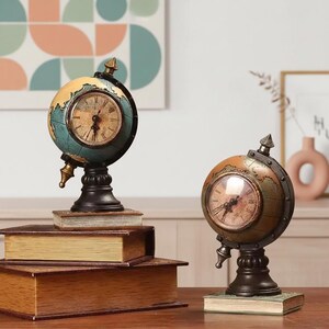 Puede incluir: Dos relojes de globo de estilo antiguo con una estética vintage. Los relojes tienen una esfera redonda con números romanos y están montados sobre una base negra. Un reloj está encima de una pila de libros. Los globos están pintados en tonos azules, dorados y marrones.