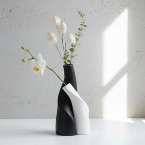 May include: A black and white ceramic vase set with a modern, intertwined design. The black vase holds dried seed pods and greenery, while a white orchid flower extends from the vase. The vases are on a white surface.
