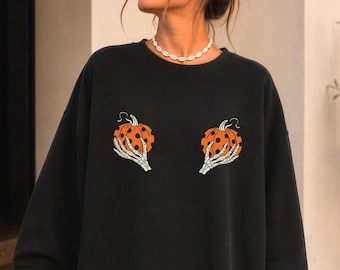 May include: A black long-sleeve crewneck sweatshirt featuring a graphic print of two skeleton hands holding orange pumpkins with black polka dots. The design is centered on the chest area of the garment, which has a relaxed fit and ribbed cuffs.