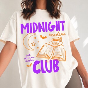 May include: An ivory t-shirt featuring a graphic print with the purple text MIDNIGHT and CLUB, and orange text reading readers and just one more chapter. The design includes an open book, a mug with a face, a crescent moon, stars, and bats.