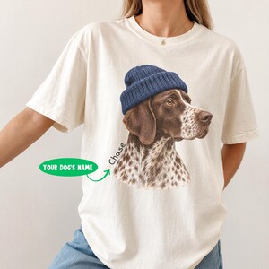 以下が含まれることがあります： アイボリーの半袖Tシャツ。青いニット帽をかぶった茶色と白の斑点模様の犬のグラフィックがプリントされています。グラフィックには「Chase」という文字と、「YOUR DOG'S NAME」と書かれた緑色の楕円形の吹き出し、および矢印が含まれています。