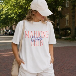May include: A white short-sleeved t-shirt featuring the red text "MAHJONG" and "CLUB" with the blue script text "Girls" in the center. The shirt is paired with white shorts, a beige shoulder bag, and a plain white baseball cap.