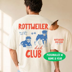 Könnte beinhalten: Ein elfenbeinfarbenes T-Shirt mit einem Rückendruck, der den roten Schriftzug ROTTWEILER DAD CLUB, zwei blaue Rottweiler-Illustrationen und den Text life is better with Max zeigt. Vorne steht ROTTWEILER DAD CLUB est. 2026.