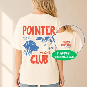 May include: An ivory t-shirt featuring a large graphic on the back with the red text POINTER MOM CLUB, a blue illustration of a pointer dog head, a full-body pointer dog, and the text life is better with Max. A circular inset shows the front chest.