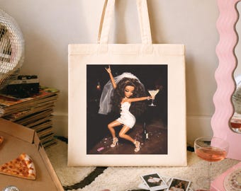 May include: A natural-colored large tote bag featuring a centered graphic print of a doll with long dark hair wearing a white dress and veil, holding a cocktail glass while dancing on a table. The text Natural large tote bag print area is visible.