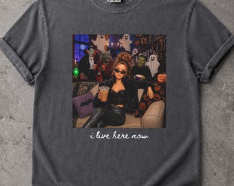 May include: A charcoal gray short-sleeve t-shirt featuring a graphic print of a person sitting on a couch surrounded by Halloween characters including ghosts, a vampire, and Frankenstein. The text "i live here now" is printed below the graphic.